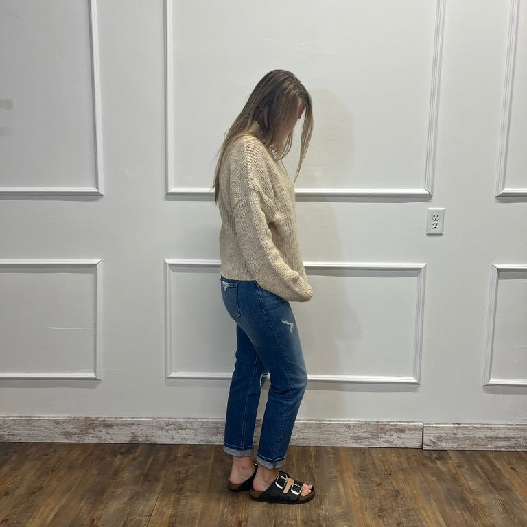 A woman stands in profile wearing a cream knit sweater, blue jeans, and black slide sandals against a white paneled wall with a wood floor.