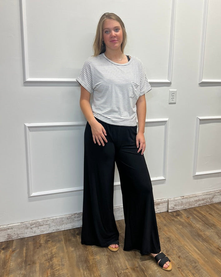 A woman modeling a short-sleeve striped t-shirt and black wide-leg pants, standing against a white paneled wall on a wood floor.