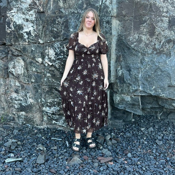 A woman stands outdoors in front of a large dark rock wall, wearing a dark brown midi dress with a white floral pattern and black sandals.