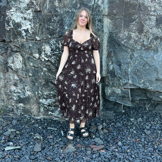 A woman stands outdoors in front of a large dark rock wall, wearing a dark brown midi dress with a white floral pattern and black sandals.