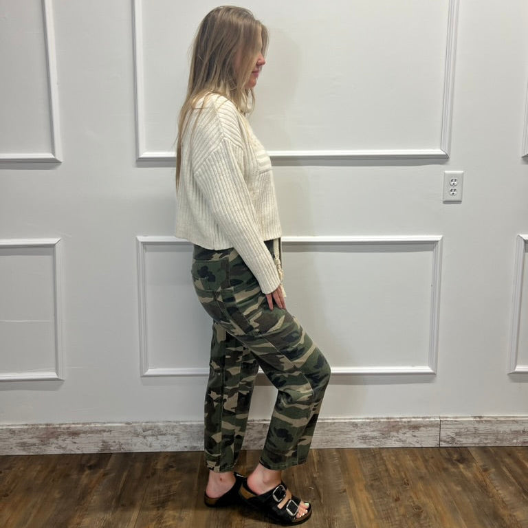A woman stands in profile wearing a cream ribbed knit cropped sweater, green camouflage cropped pants, and black slide sandals, set against a white paneled wall and wood floor.