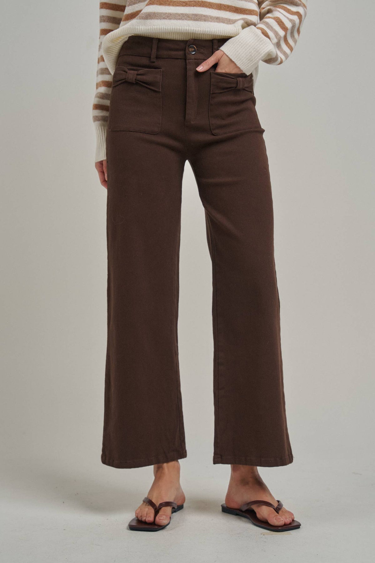 A person wearing dark brown wide-leg cropped pants with a striped cream and brown sweater, paired with black flip-flop sandals, photographed against a light gray studio background.