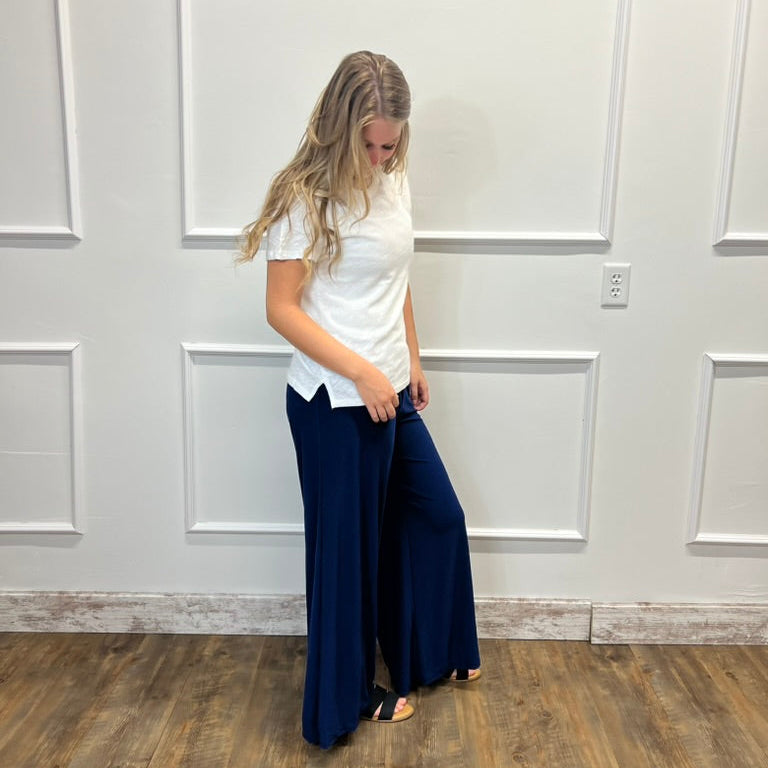 A woman stands in a room with white paneled walls and a wood floor, wearing a white short-sleeve top and dark navy wide-leg pants.