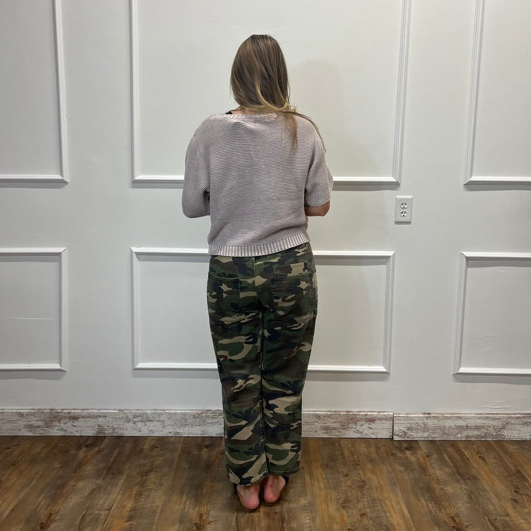 A woman viewed from behind wearing a light gray knit sweater and camouflage pants, standing on a wood floor against a white paneled wall.