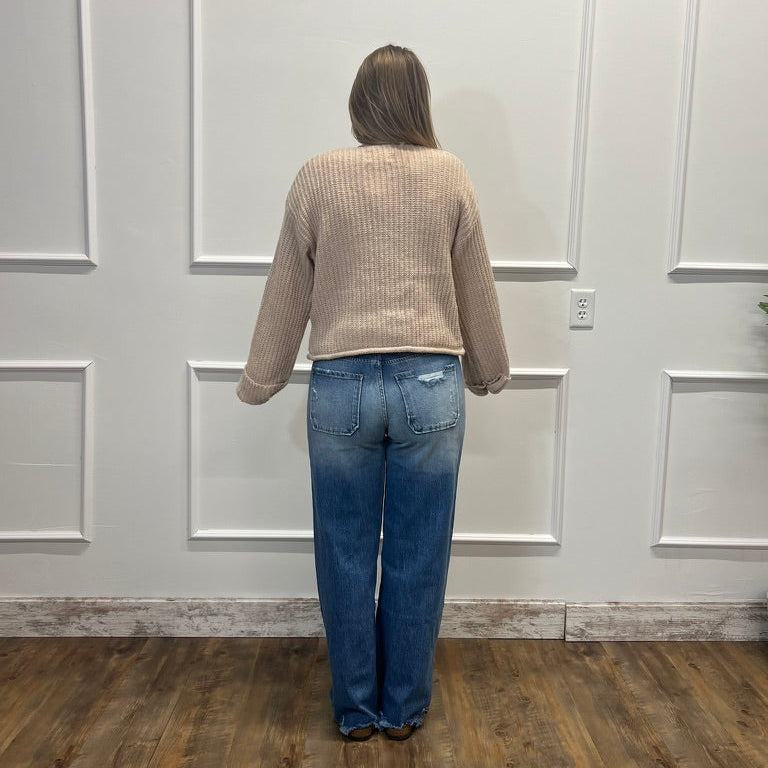 A person viewed from behind wearing a beige ribbed knit sweater and blue jeans, standing on a wood floor against a white paneled wall.