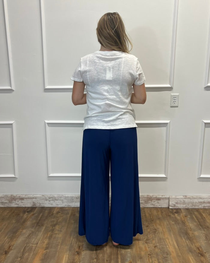 A woman viewed from behind wearing a white short-sleeve top and navy blue wide-leg pants, standing on a wood floor against a white paneled wall.