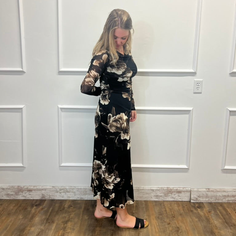 A woman modeling a long-sleeve black maxi dress with a beige and white floral pattern, standing against a white paneled wall on a wood floor.