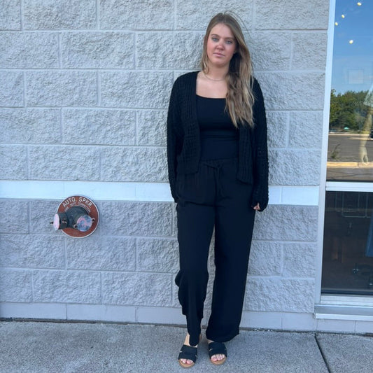 A woman stands outdoors against a light gray stone wall wearing a black outfit consisting of a tank top, wide-leg pants, and a black cardigan, paired with black slide sandals.