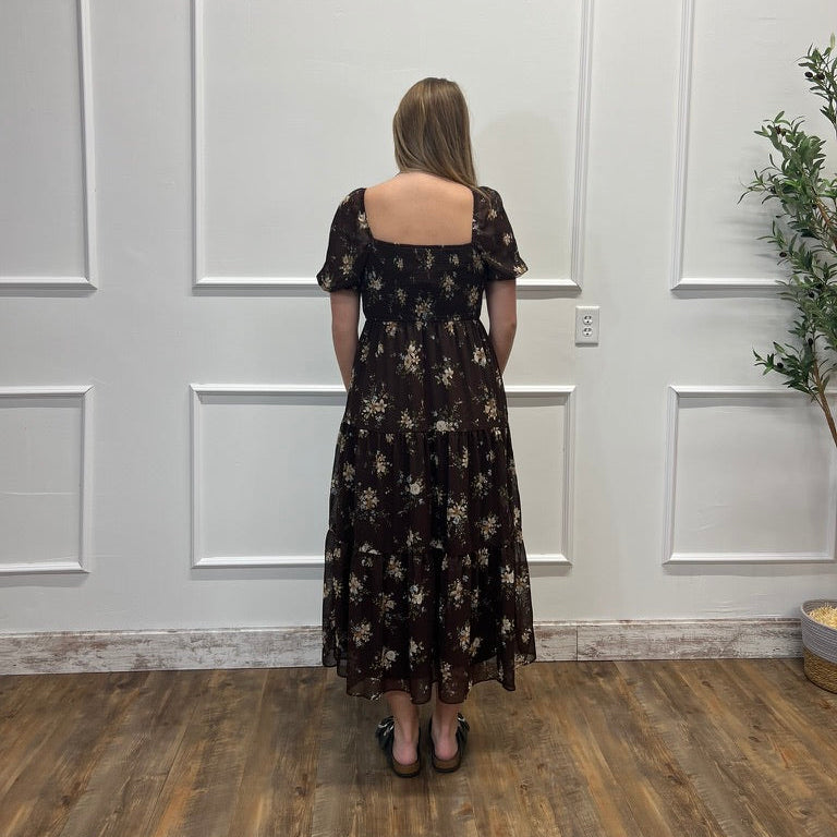 A woman stands with her back to the camera wearing a dark brown midi-length dress with a beige floral pattern, short puff sleeves, and a square neckline. She is standing in front of a white paneled wall on a wooden floor.