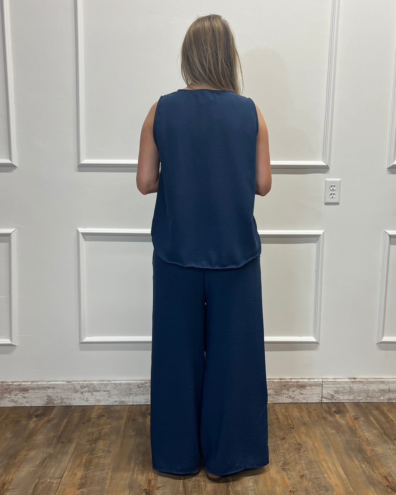 A woman viewed from behind wearing a sleeveless navy blue top and matching wide-leg navy pants, standing in front of a white paneled wall on a wood floor.