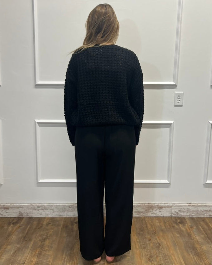 A woman stands with her back to the camera wearing a black textured knit sweater and black wide-leg pants in front of a white paneled wall with a wood floor.