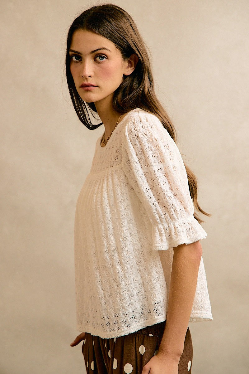 A woman wearing a cream-colored eyelet lace short-sleeve blouse with a gathered waist, paired with brown polka-dot pants, posing against a textured beige studio backdrop.