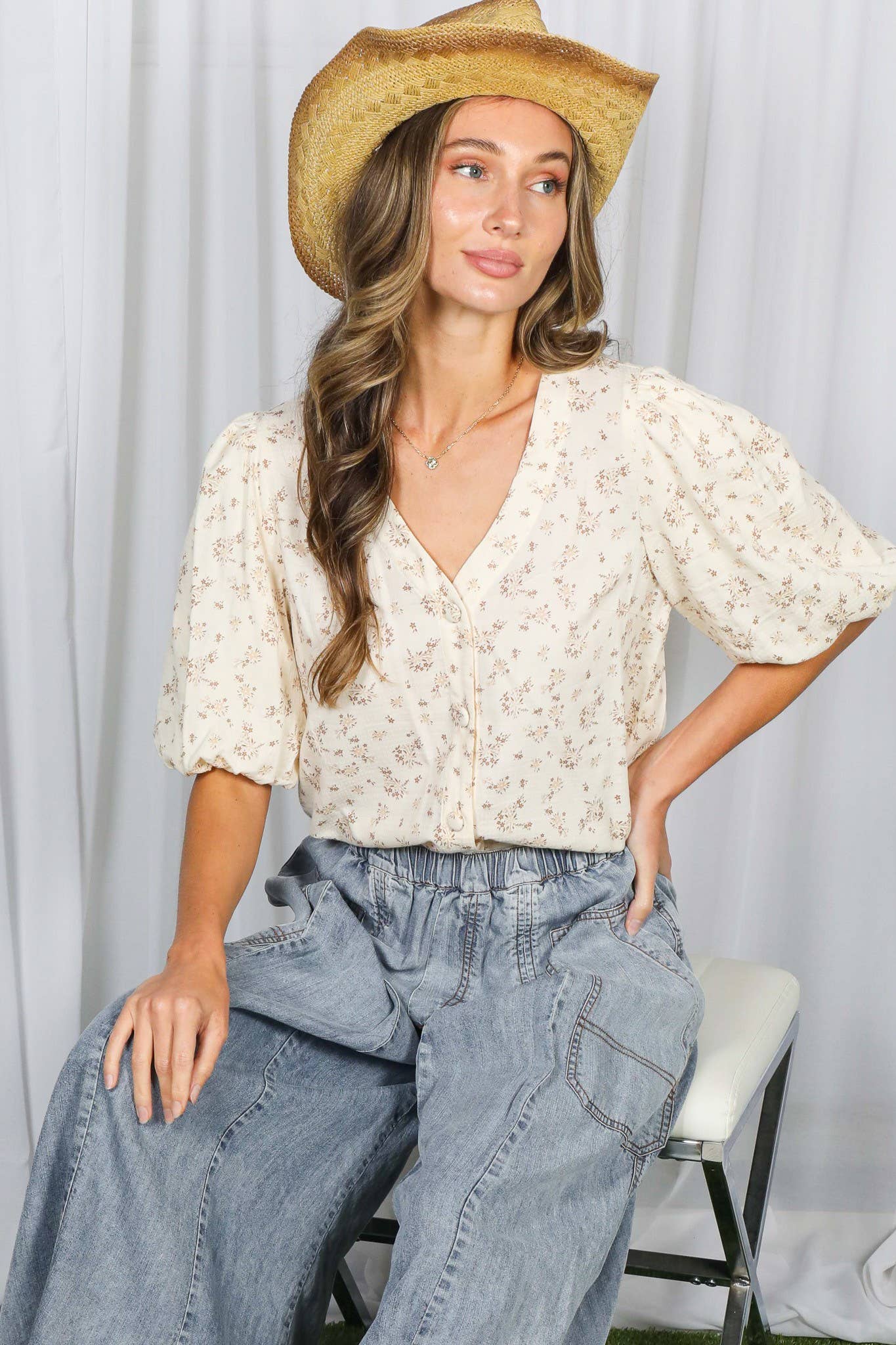 Woman wearing a beige blouse and blue jeans with a straw hat against a white curtain background