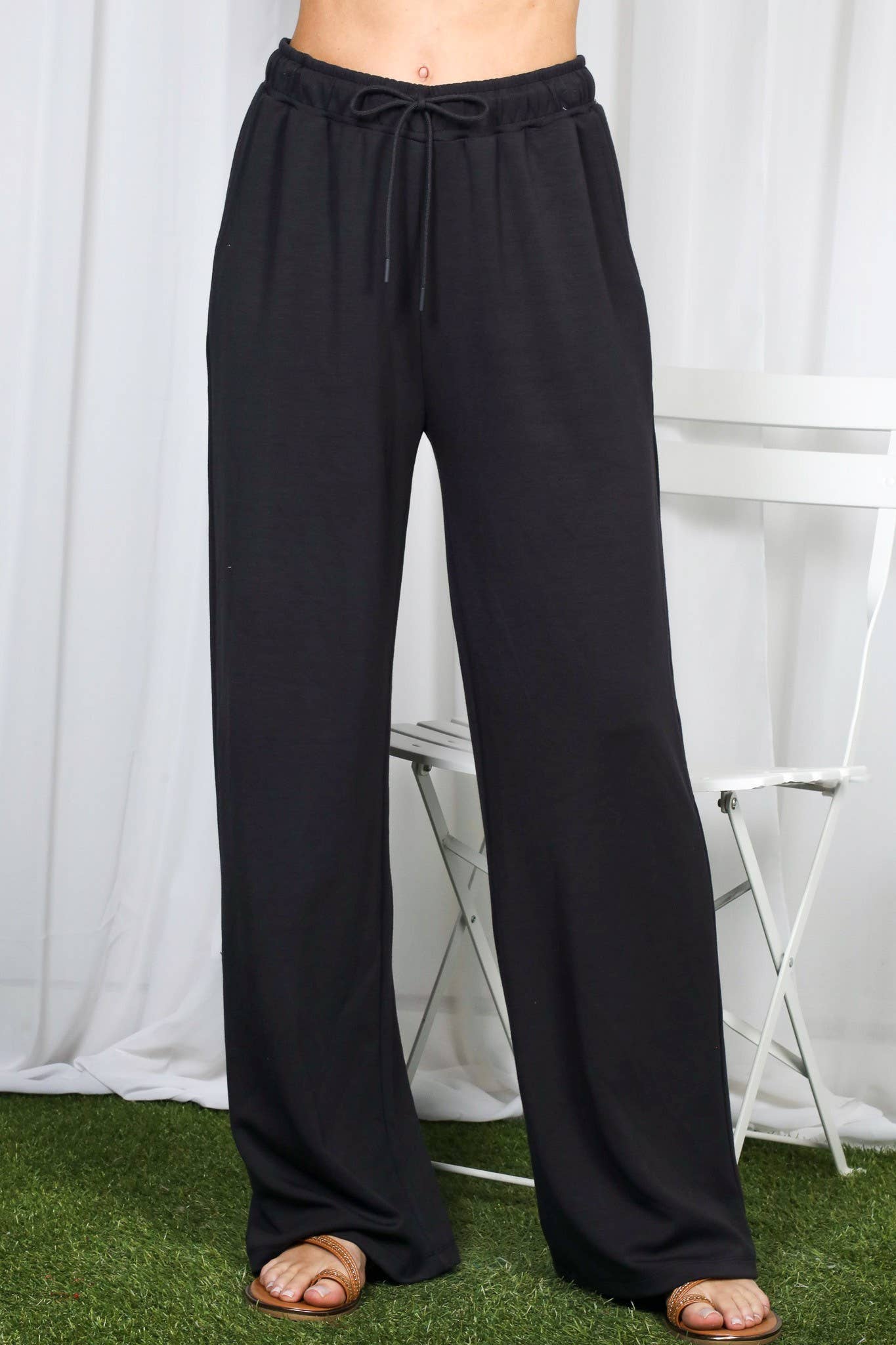 Black wide-leg drawstring pants shown on a model from the waist down, standing on artificial green turf in front of a white curtain backdrop.