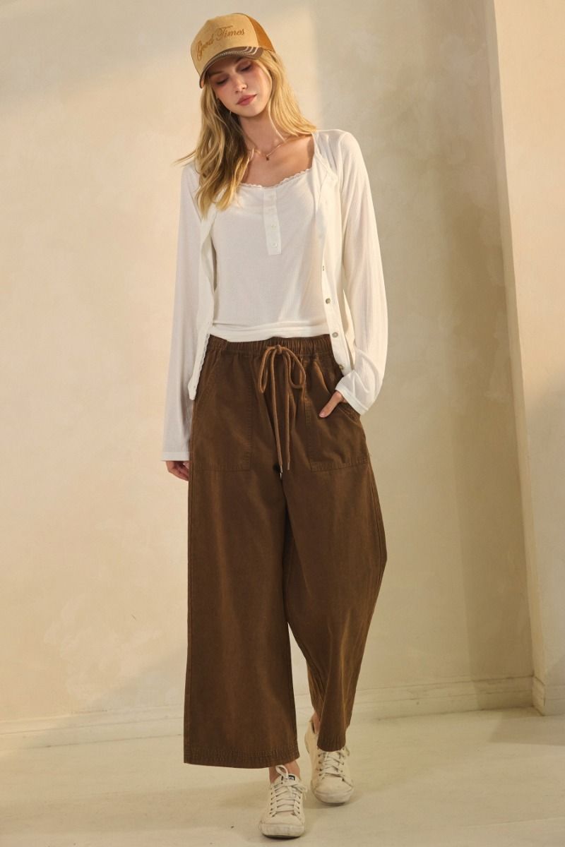 Woman wearing a white blouse, brown wide-leg pants, and a beige cap against a plain background