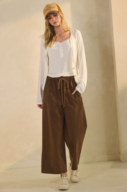 Woman wearing a white blouse, brown wide-leg pants, and a beige cap against a plain background