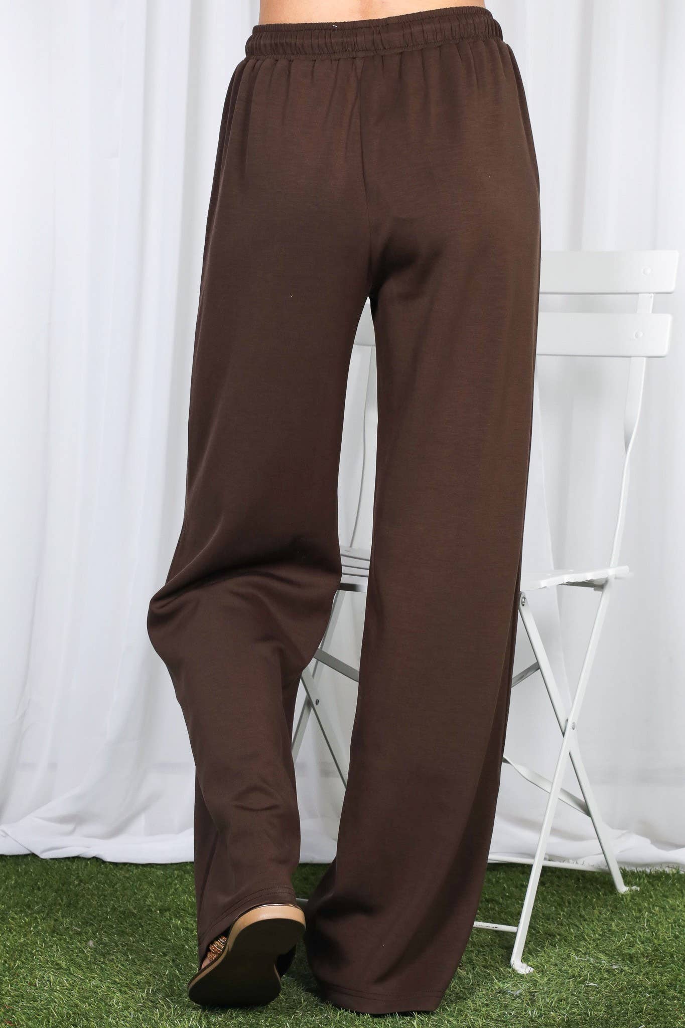 Brown wide-leg lounge pants shown on a model from the waist down, standing on artificial green grass in front of a white curtain backdrop with a white folding chair.
