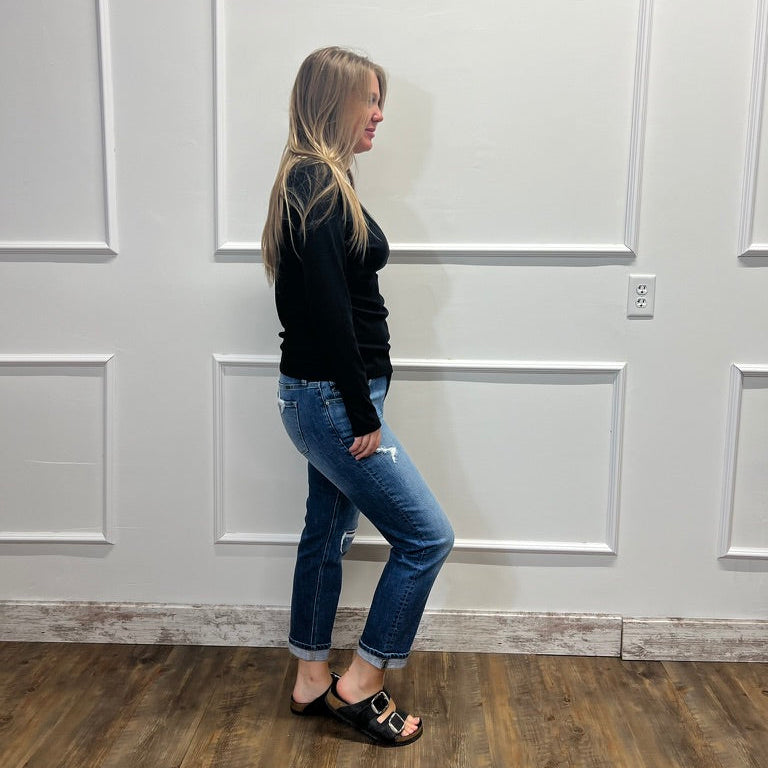 A woman stands in profile wearing a black long-sleeve top, blue cropped jeans, and black slide sandals against a white paneled wall with wood-look flooring.