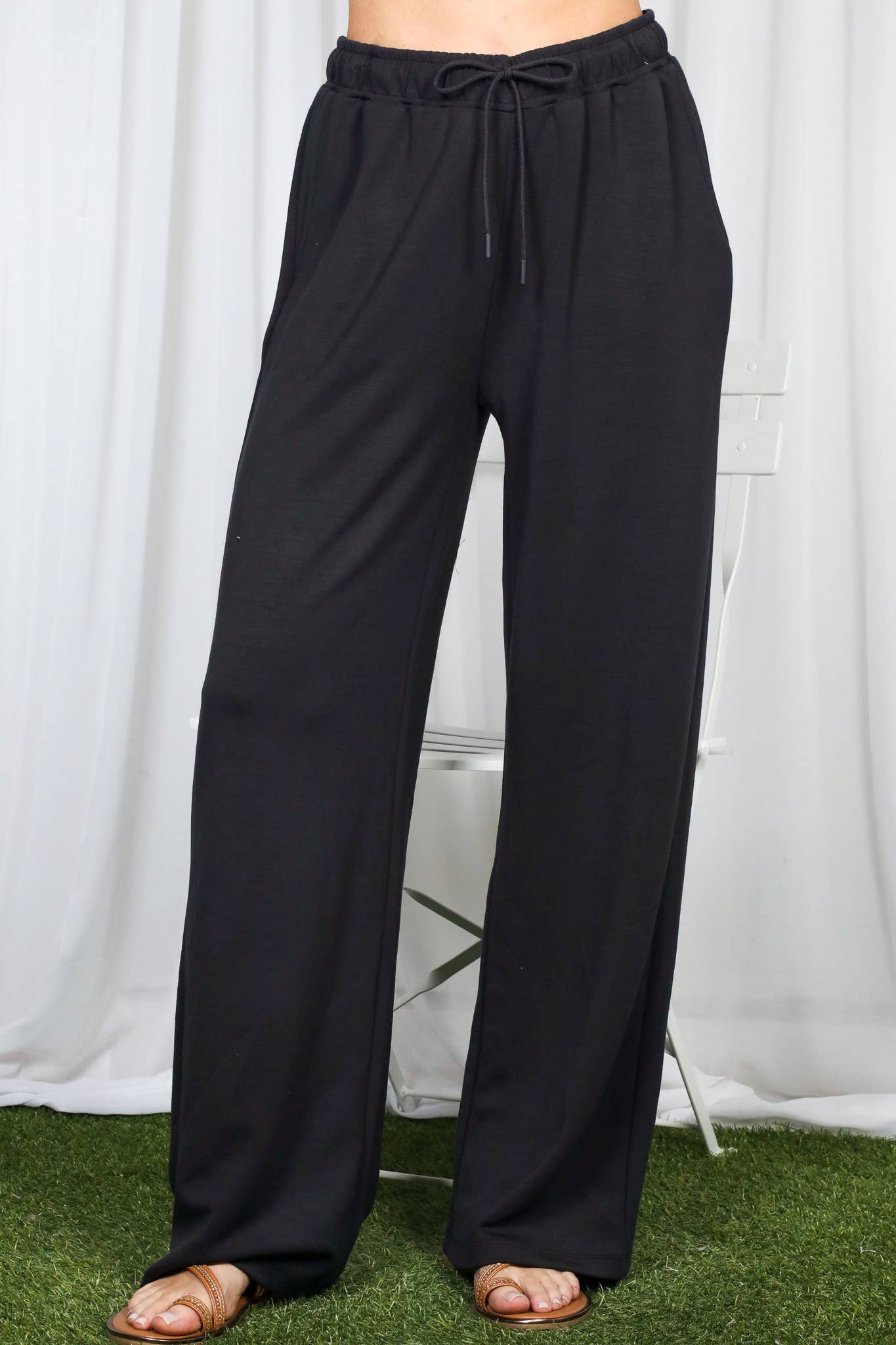 Black wide-leg drawstring pants shown on a model from the waist down, standing on artificial green grass in front of a white curtain backdrop.