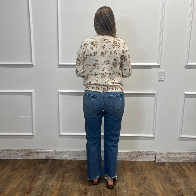 A woman viewed from behind wearing a cream floral long-sleeve top and blue jeans, standing on a wood-look floor against a white paneled wall.