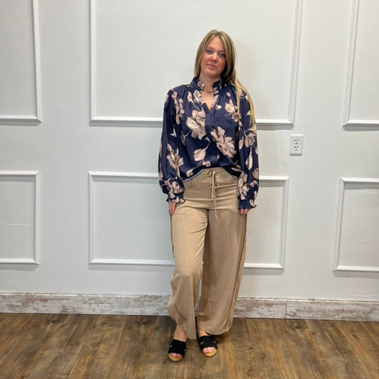 A woman stands in a room with white paneled walls and a wood floor, wearing a navy blue floral long-sleeve blouse and beige wide-leg pants.