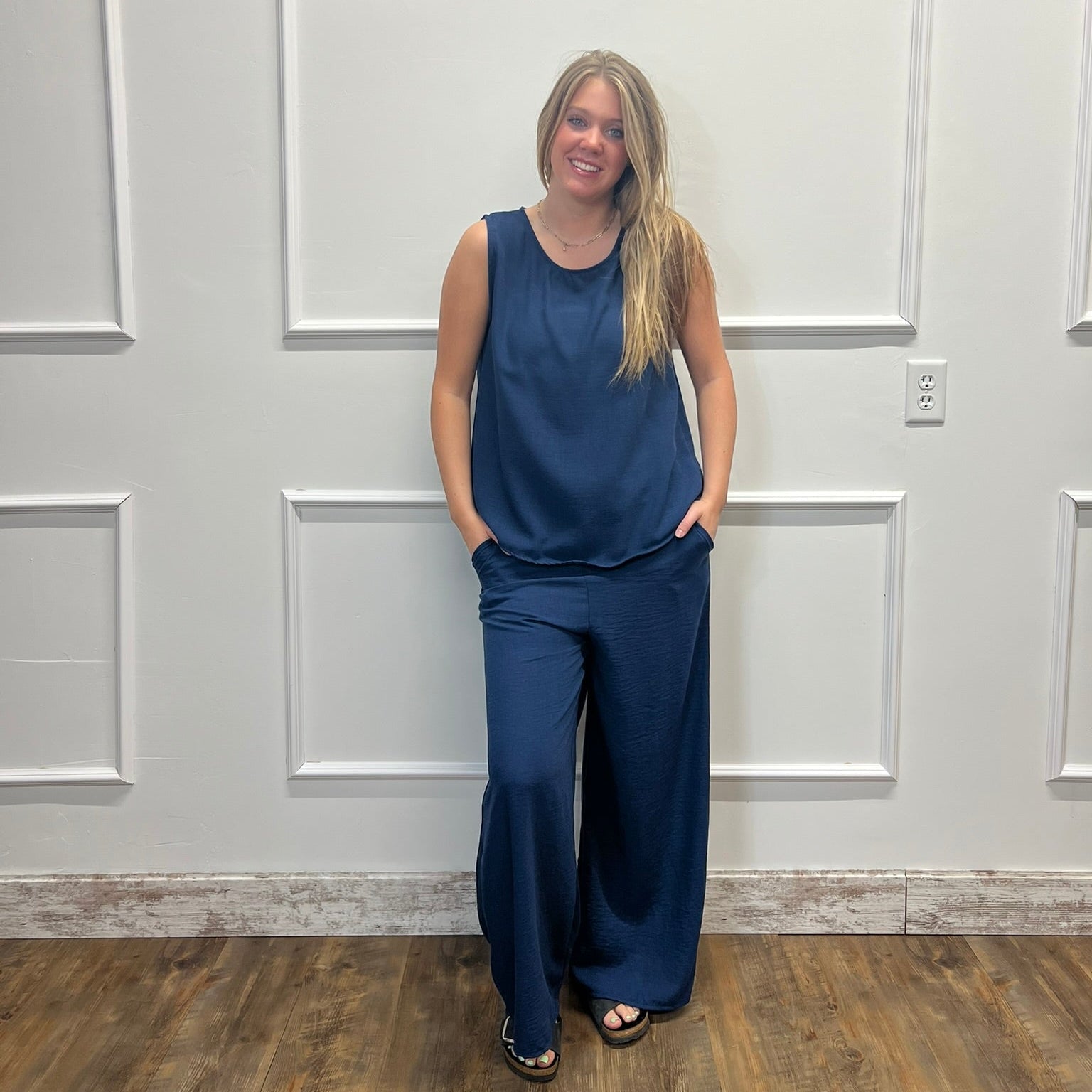 A woman modeling a navy blue sleeveless top and matching wide-leg pants, standing against a white paneled wall with wood flooring.