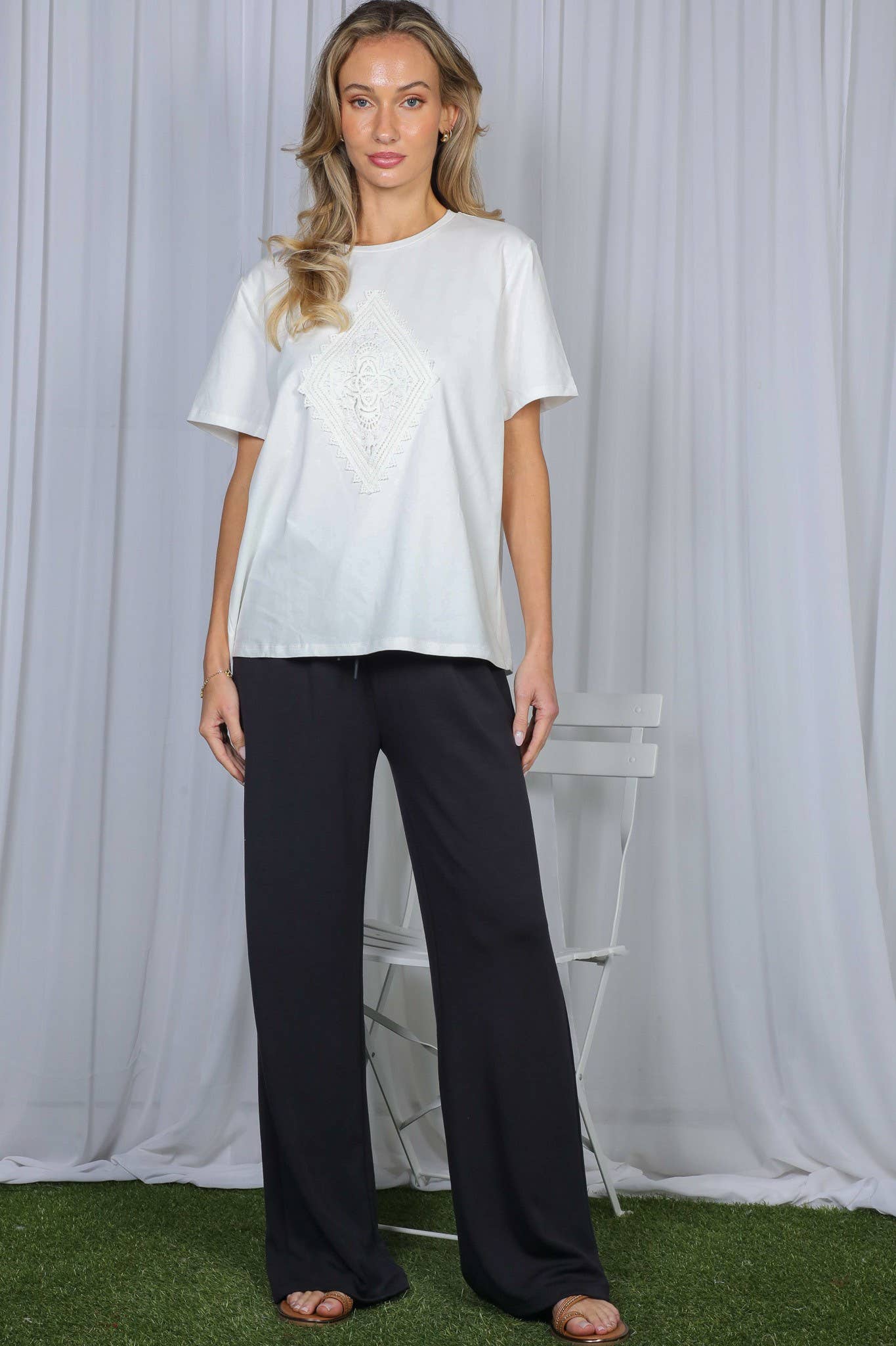 A woman modeling a white short-sleeve t-shirt with a textured chest design and black wide-leg pants, standing in front of a white draped curtain backdrop with artificial green grass at the bottom.