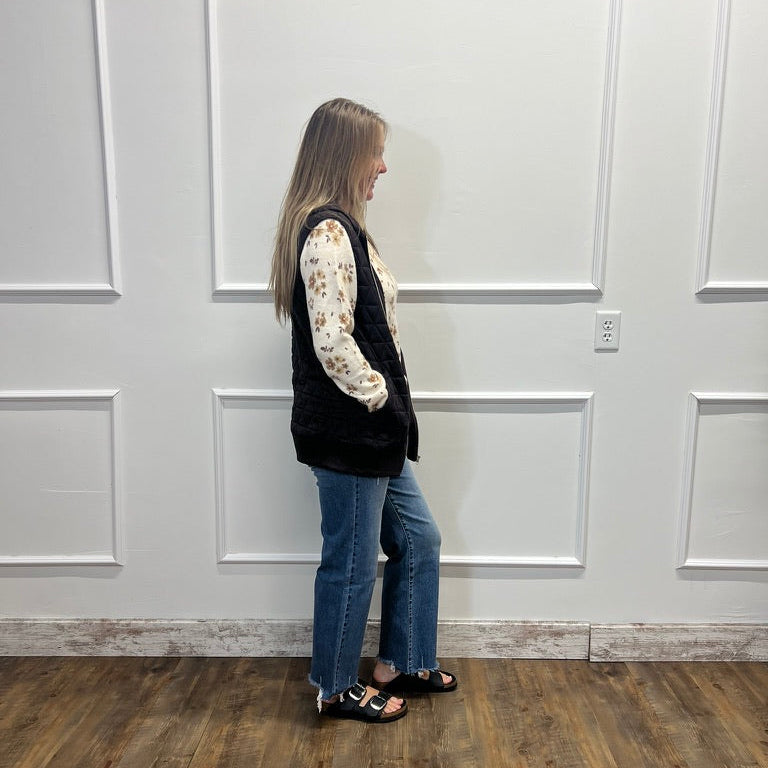 A woman stands in profile wearing a black sleeveless vest over a cream floral long-sleeve shirt, blue jeans, and black sandals, standing against a white paneled wall on a wood-look floor.