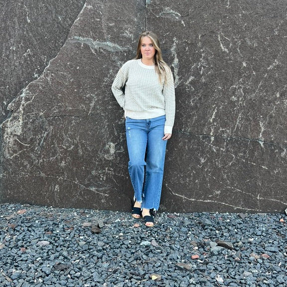 A woman stands in front of a large dark rock formation outdoors, wearing a light beige long-sleeve top, blue jeans, and black sandals on a gravel ground.