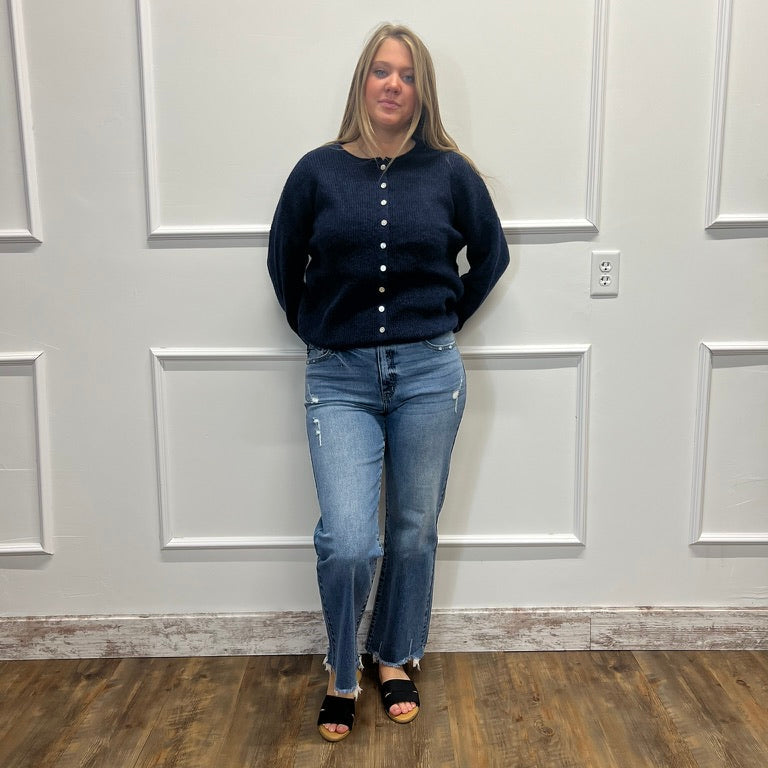 A woman stands in a room with white paneled walls and a wood floor, wearing a dark navy blue button-up cardigan, blue jeans, and black sandals.
