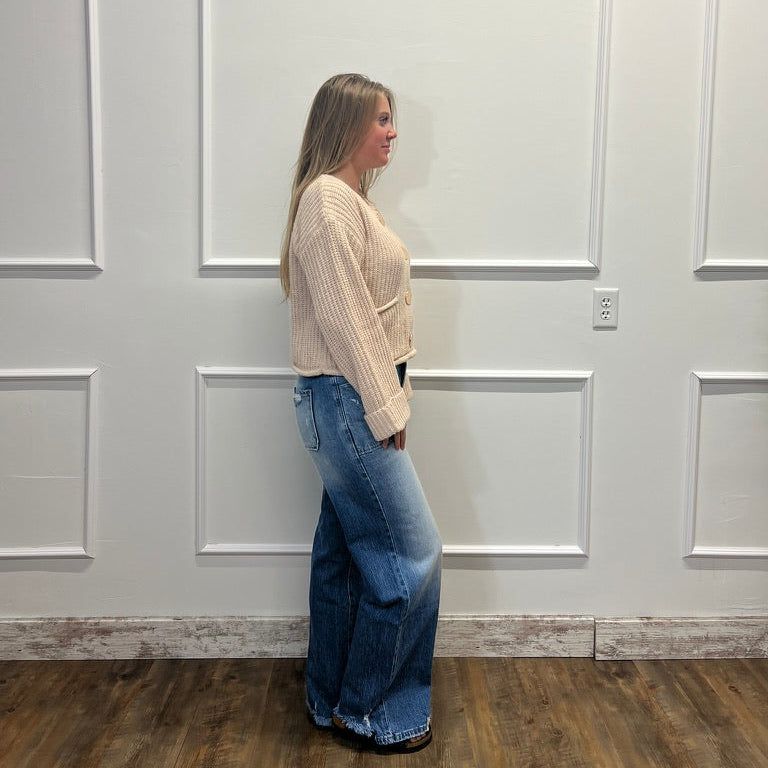 A woman stands in profile wearing a cream ribbed knit cropped sweater and wide-leg blue jeans against a white paneled wall with a wood floor.