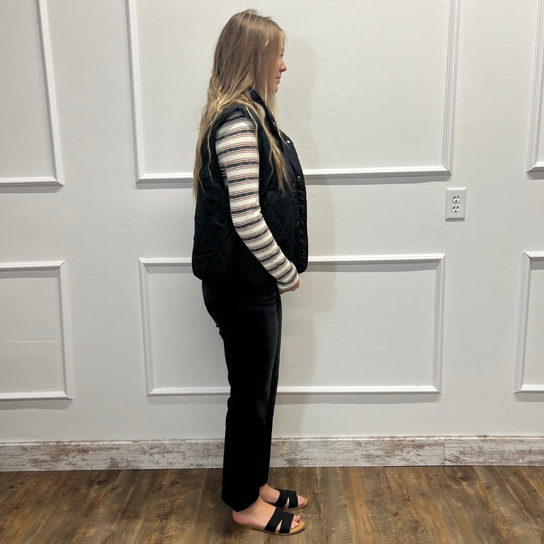 A woman stands in profile wearing a black quilted sleeveless vest over a black and white striped long-sleeve shirt, black pants, and black slide sandals, standing in front of a white paneled wall on a wood floor.