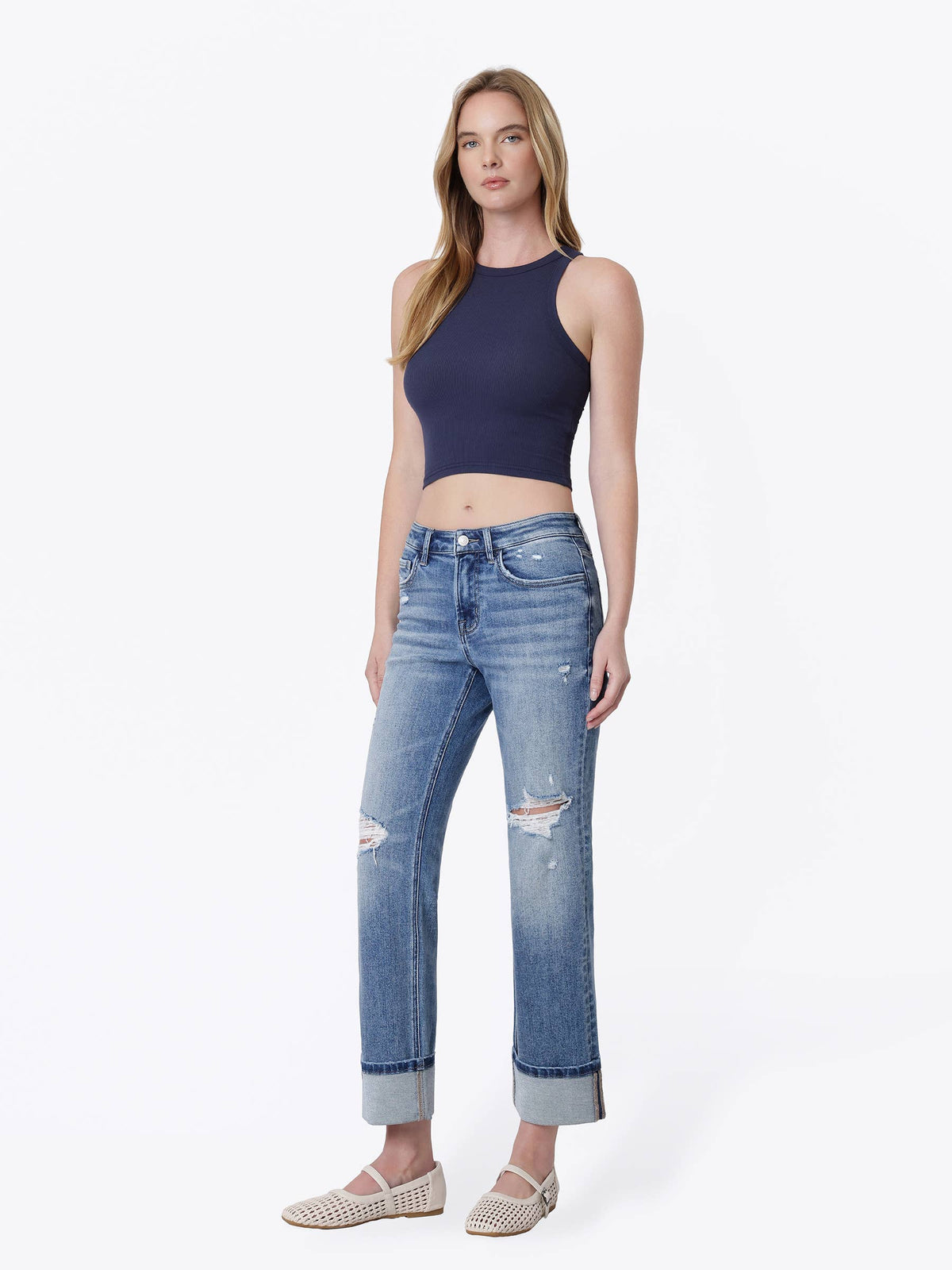 A woman modeling a dark navy blue ribbed halter crop top and light-wash blue distressed jeans with rolled cuffs, paired with beige woven flat shoes, against a plain white studio background.