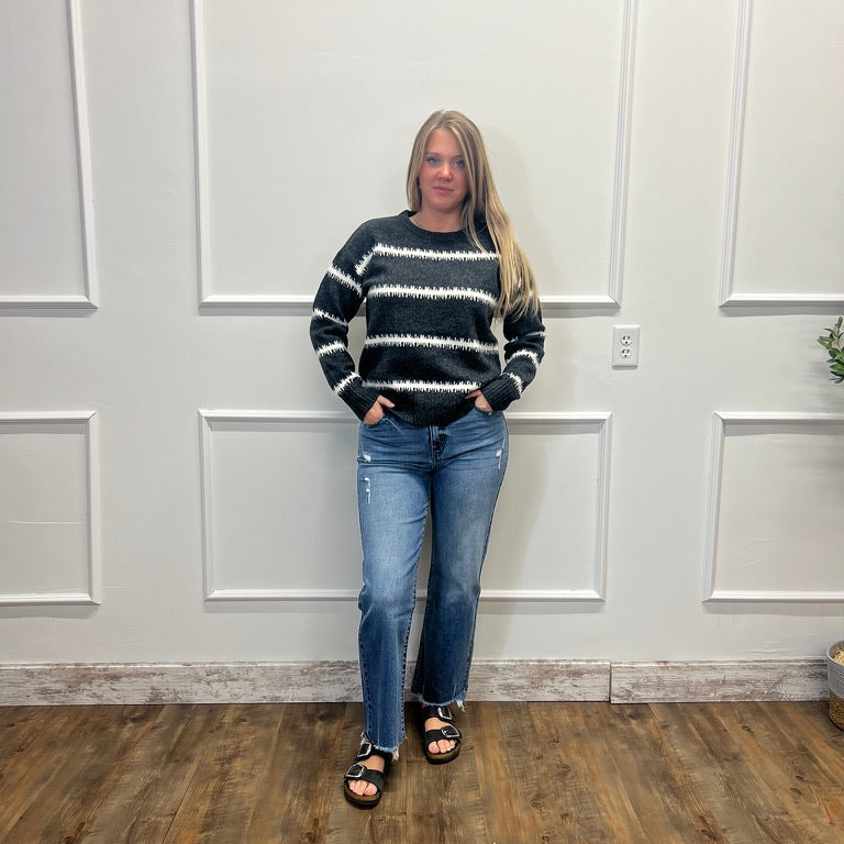 A woman stands indoors against a white paneled wall wearing a black and white horizontally striped knit sweater, blue jeans, and black sandals.