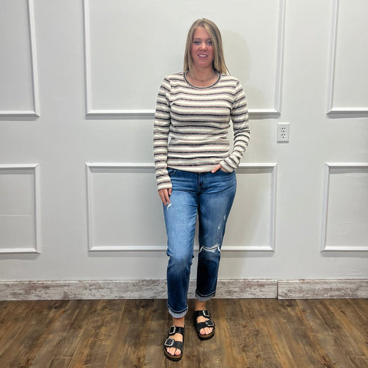 A woman stands in a room with white paneled walls and a wood floor, wearing a beige and white striped long-sleeve knit top, blue jeans, and black slide sandals.