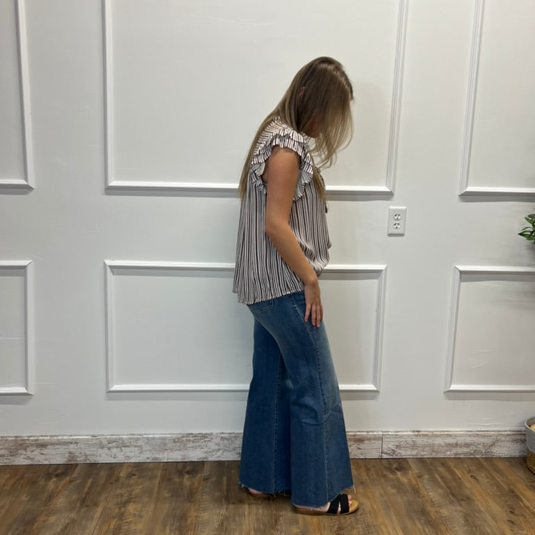 A woman stands in profile wearing a sleeveless ruffled top with vertical stripes in beige and white, paired with wide-leg blue jeans and black sandals, set against a white paneled wall and wood floor.
