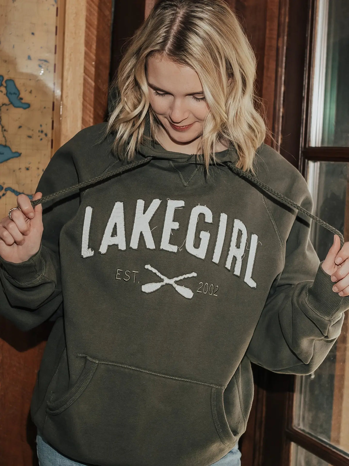 A woman wearing an olive green hoodie with 'LAKEGIRL EST. 2002' printed in white across the chest, holding the drawstrings open.