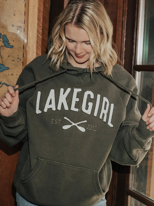 A woman wearing an olive green hoodie with 'LAKEGIRL EST. 2002' printed in white across the chest, holding the drawstrings open.