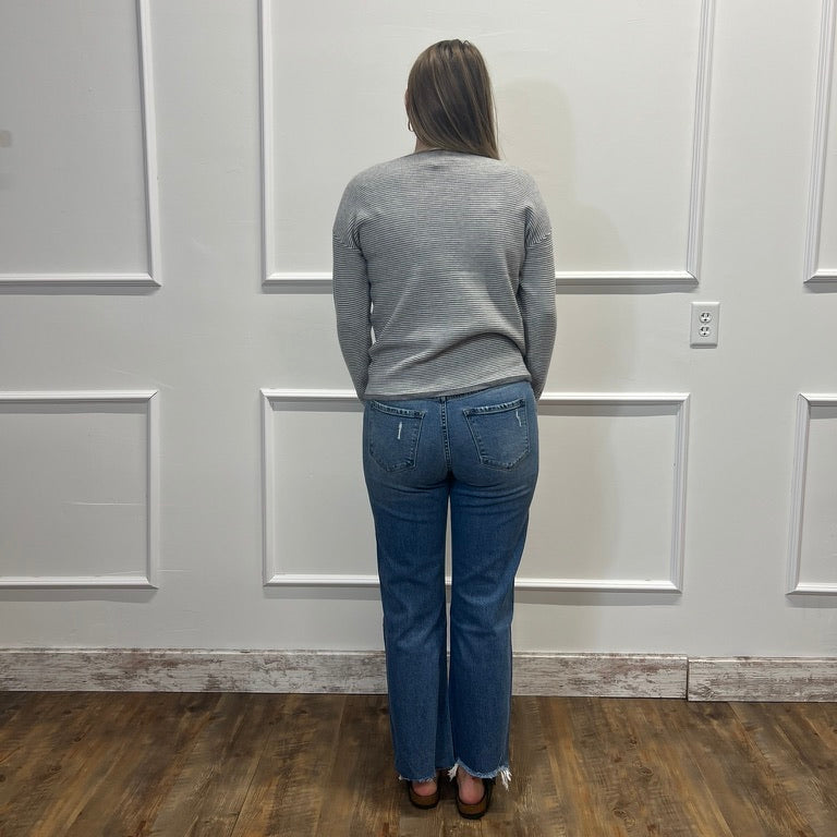 A woman viewed from behind wearing a gray long-sleeve knit sweater and blue jeans, standing on a wood floor against a white paneled wall.