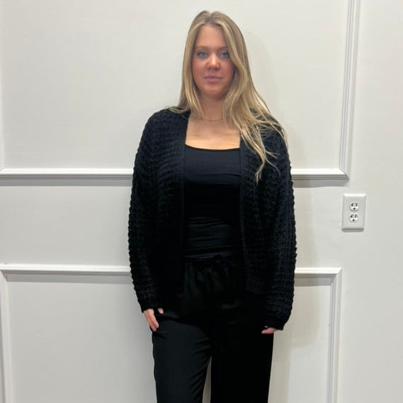 A woman stands indoors wearing a black textured cardigan, black top, black pants, and black slide sandals against a white paneled wall with wood flooring.
