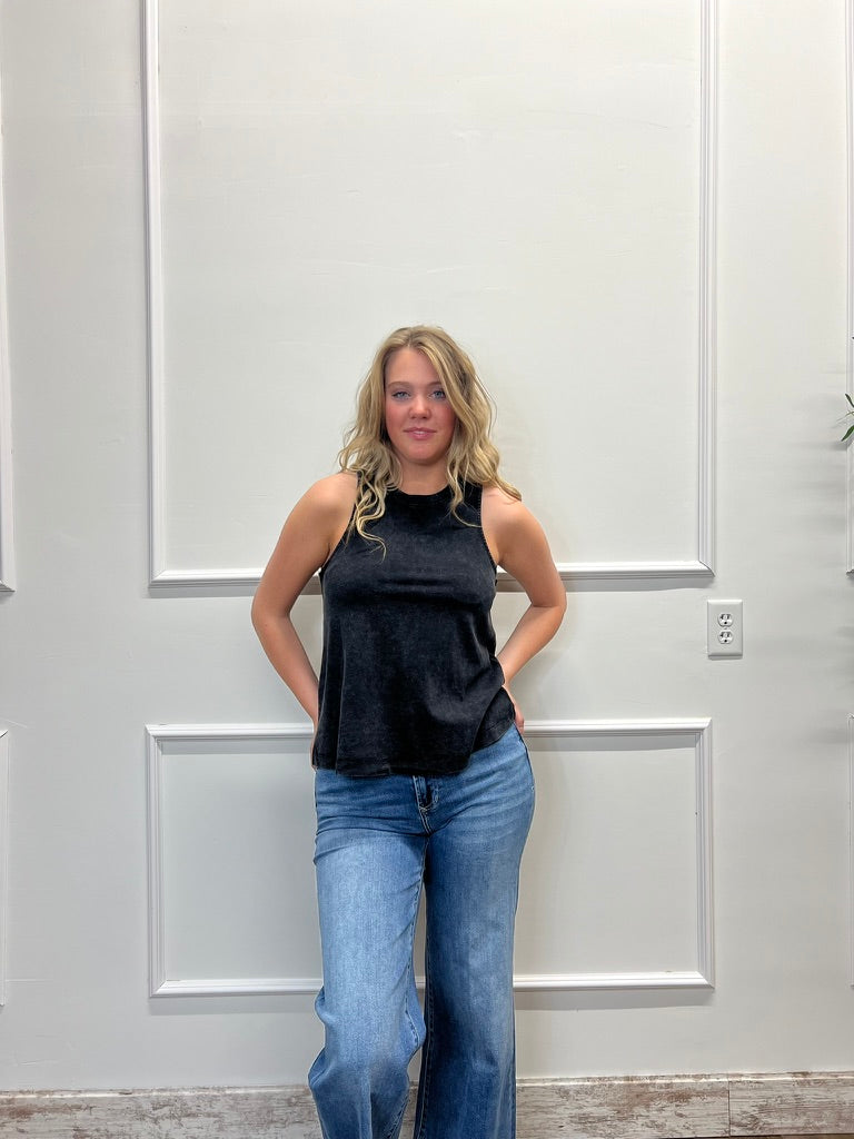Woman wearing a black sleeveless top and blue jeans standing against a white paneled wall.