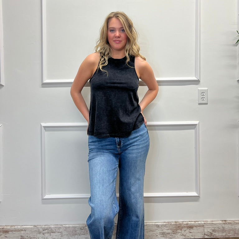 Woman wearing a black sleeveless top and blue jeans standing against a white paneled wall.