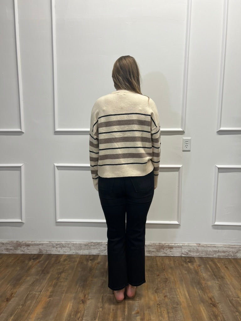A woman viewed from behind wearing a cream knit sweater with brown and black horizontal stripes, paired with black pants, standing in front of a white paneled wall on a wood floor.