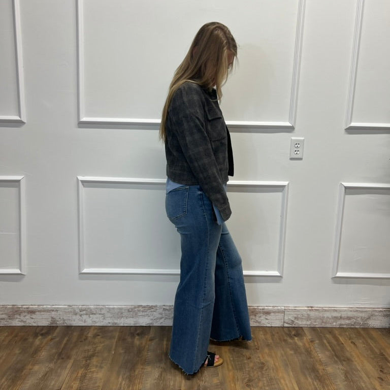 A woman stands in profile wearing a dark gray plaid flannel shirt and blue flare jeans, standing on a wood floor against a white paneled wall.