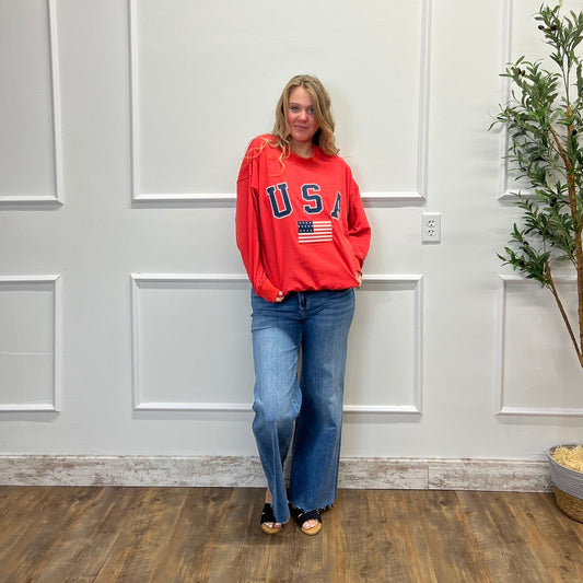 Woman wearing a red 'USA' sweatshirt and blue jeans standing in a room with a white paneled wall and wooden floor.