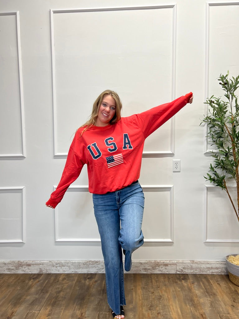 Person wearing a red 'USA' sweatshirt with an American flag graphic, standing in a room with a white paneled wall and wooden floor.