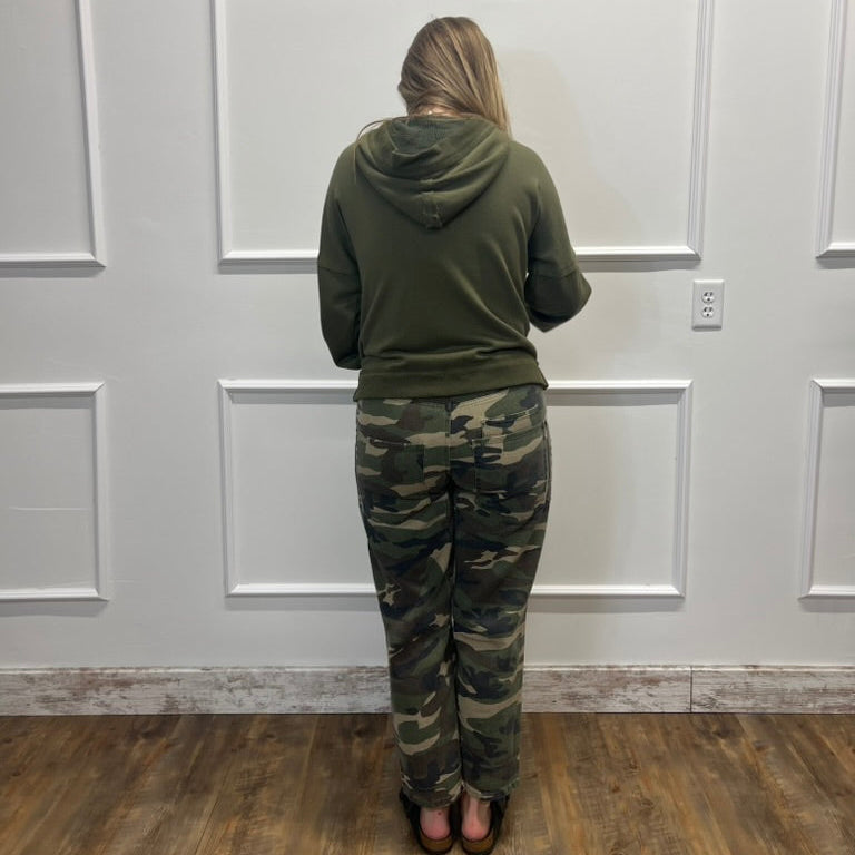 A woman viewed from behind wearing an olive green hoodie and camouflage pants, standing against a white paneled wall on a wood floor.