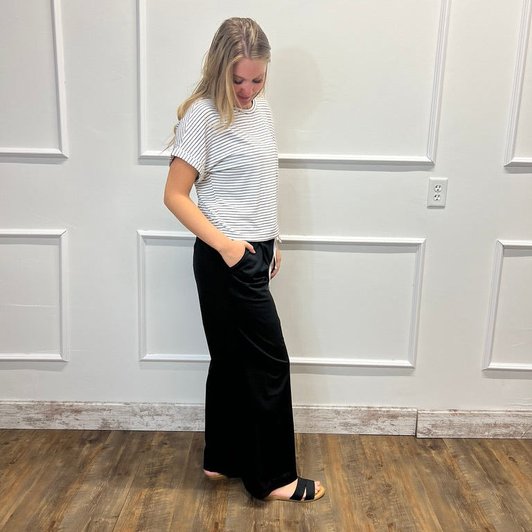 A woman stands in profile wearing a white short-sleeve t-shirt with thin black horizontal stripes and black wide-leg pants, standing on a wood-look floor against a white paneled wall.