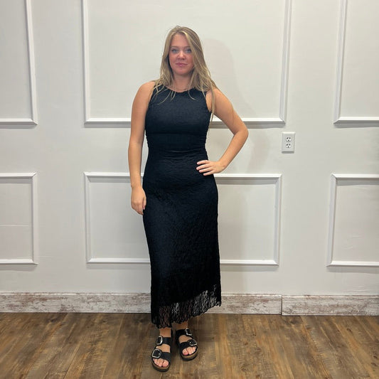 A woman stands in a black sleeveless maxi dress with a textured lace-like pattern and a sheer fringe hem, paired with black sandals, posing against a white paneled wall on a wood floor.