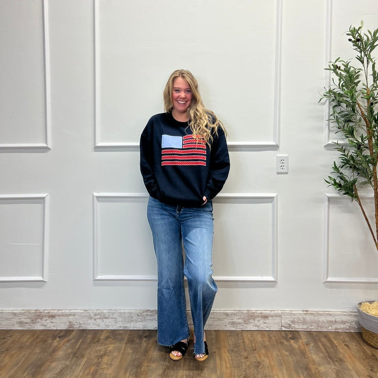 Woman wearing a black sweater with a colorful flag design and blue jeans standing in a room with white paneled walls and a plant.